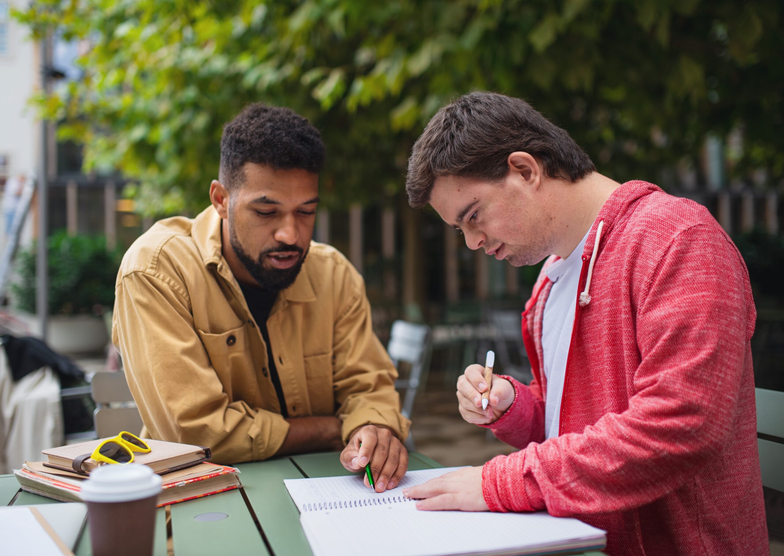 A young man with Down syndrome with his mentoring friend sitting outdoors in cafe and studying.