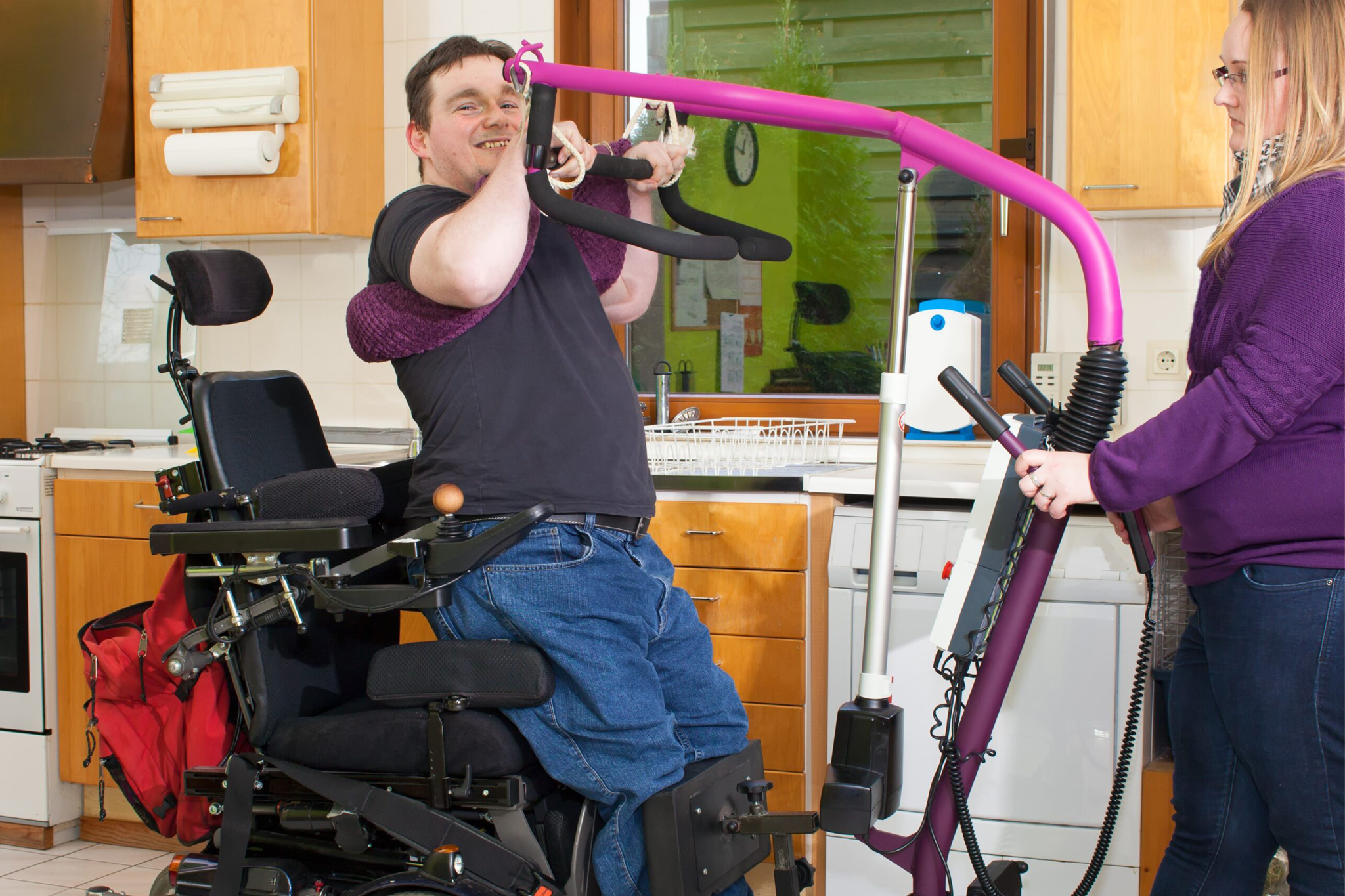 Therapist helping a spastic young man with infantile cerebral palsy caused by a complicated birth to maneuver himself into a multifunctional wheelchair with a patient lift.